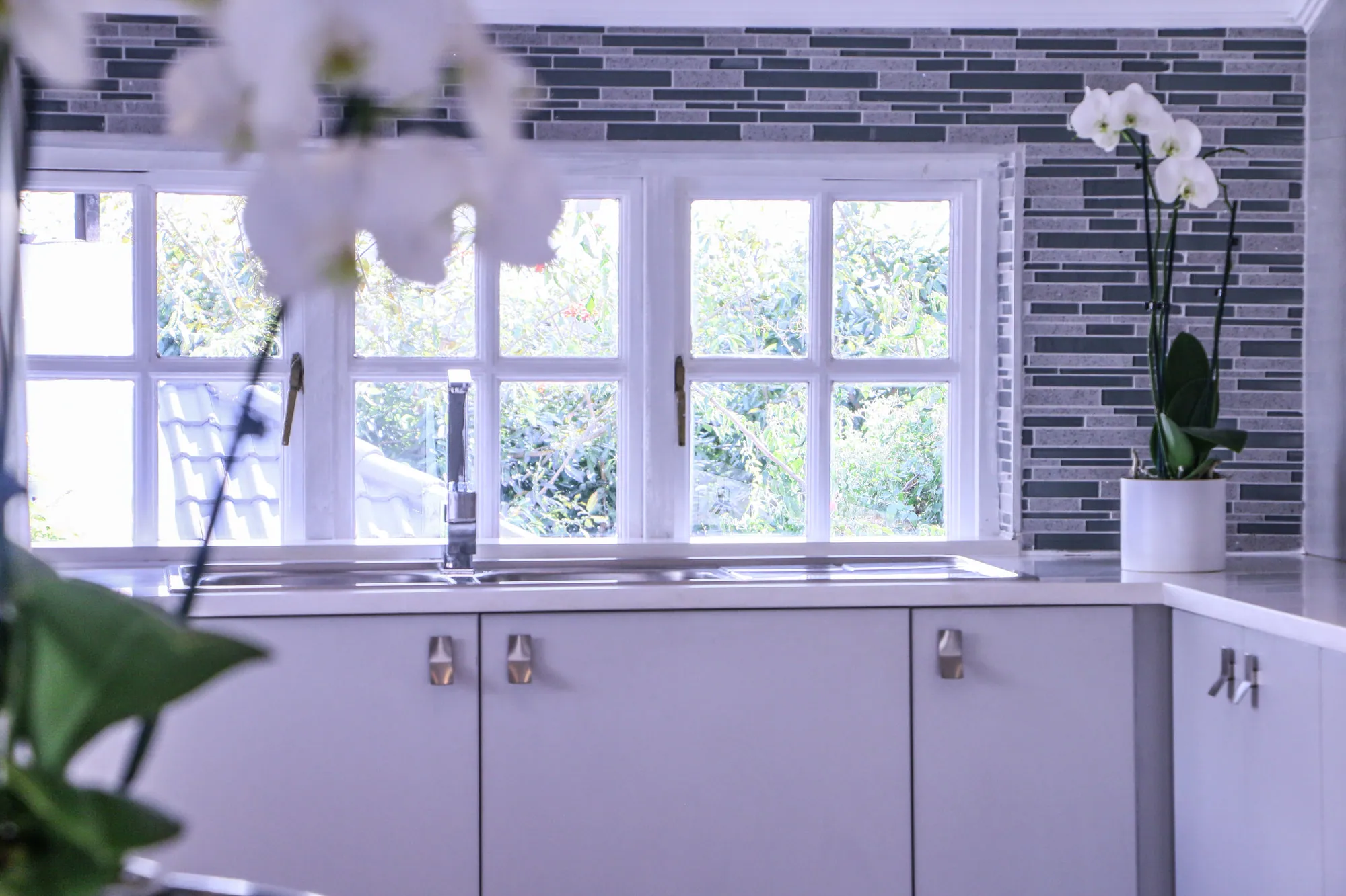 White kitchen sink run beneath a window with mosaic-tiled feature wall