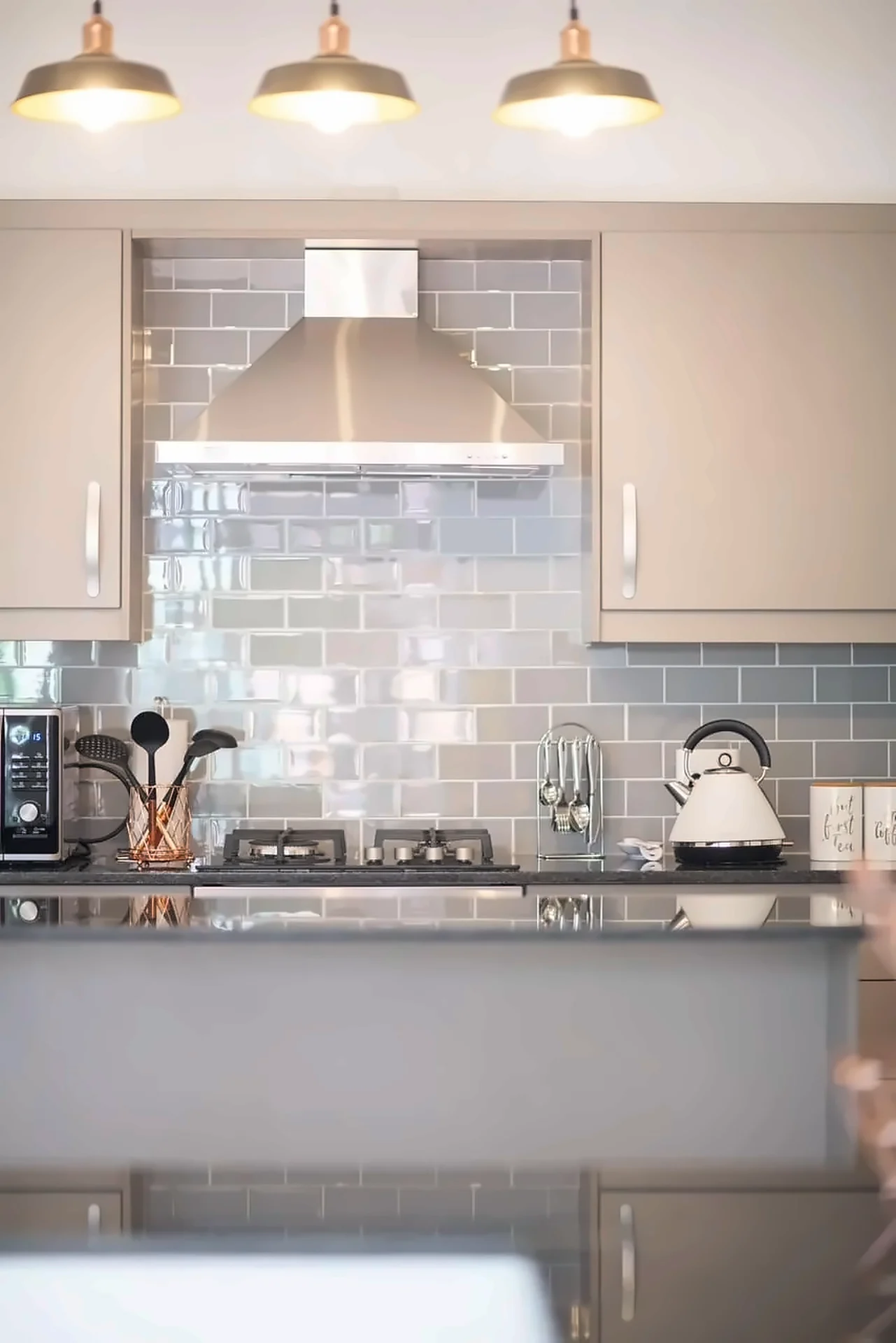Grey cabinets with blue-grey subway tiles and stainless extractor under three pendants